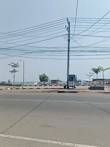 A motorcycle with a rider wearing a white shirt and a white helmet is traveling from left to right across a paved road. The road has white lane markings. In the background, there is a body of water, possibly the ocean, with boats visible. There are also trees, small buildings, and utility poles with many wires overhead. A small booth or kiosk is positioned near a utility pole on the right side of the road. The sky is clear and bright. The scene appears to be a coastal road on a sunny day.