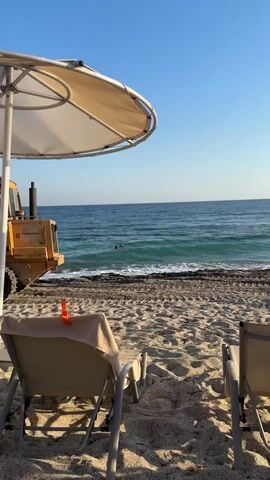 A yellow bulldozer is parked on a sandy beach next to the ocean. There are two lounge chairs in the foreground, one with a towel draped over it and a pair of sunglasses. The sky is clear and blue.