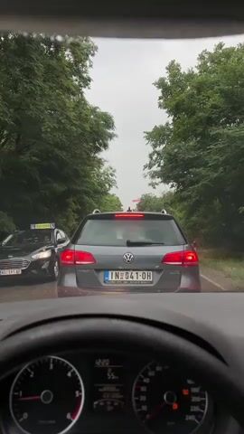 The video is captured from inside a car, looking out through the windshield. The car appears to be in traffic, with a grey Volkswagen wagon directly in front. The Volkswagen has a license plate that reads IN 041 OH. The scene is outdoors, with lush green trees lining both sides of the road, suggesting a rural or suburban setting. The sky is overcast and appears to be grey, indicating possibly cloudy or rainy weather. The dashboard of the car is partially visible in the foreground, showing the speedometer with a reading of approximately 55, and other indicators. A red object, possibly a light or a sign, is visible in the distance above the trees. Another car, partially visible on the left, has text on its side that includes "ČAK". The overall impression is a moment captured in a traffic jam on a road surrounded by nature.