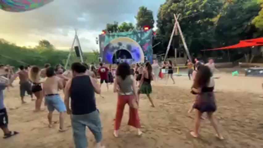 A crowd of people are dancing on a beach, facing a stage with a DJ booth. The stage is decorated with psychedelic patterns and lit with blue and white lights. The crowd is diverse in age and dress, and most are moving to the music. A colorful psychedelic canopy hangs above the dancing area. The environment includes trees, simple structures like wooden tripods, and a red umbrella. The sky is overcast, but with a glimpse of the setting sun, providing ambient lighting. The scene feels energetic, carefree, and indicative of an outdoor music festival or party.