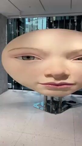 The video shows two robotic faces on display. The first face, close to the camera, is oval-shaped and large. It has realistic features, including freckles, light pink lips, and light-colored eyes. The second face, smaller and further back, is more traditionally human in shape. It has a neutral expression. Both faces are mounted on metal stands. The background shows a modern interior with a geometric pattern on the walls and large windows.