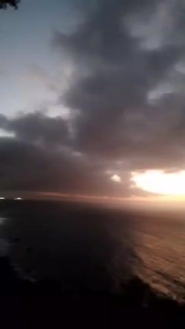 A short video clip shows a view of the ocean at sunset. The sky is filled with dark, cloudy formations. The sun is setting on the horizon, casting a warm orange and yellow glow over the water. The reflection of the sun creates a shimmering path across the ocean's surface. In the foreground, the silhouettes of trees and foliage are visible against the fading light. The video is grainy and appears to be recorded with a low-resolution camera.