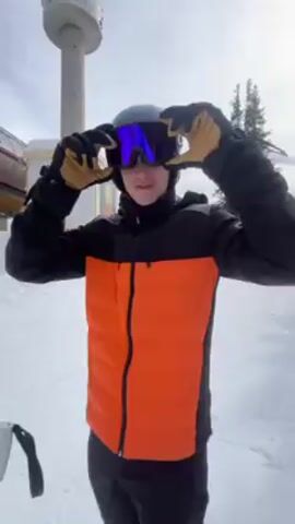 The video shows a person standing in the snow, wearing a black and orange winter coat, a grey helmet, and purple ski goggles. They are wearing gloves and make a wide gesture with their hands, as if indicating the vastness of the scene. Behind them is a snow-covered landscape, a small building, a ski lift, and a tall, tower-like structure.