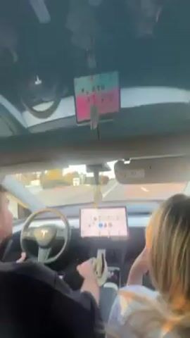 Inside a white Tesla, a man drives and a woman sits in the passenger seat. The dashboard screen is visible. A decorative object hangs from the rearview mirror. A phone sits in the center console, next to what looks like a beige hat. The car appears to be moving on a highway or major road. A camera in the sunroof displays footage in reverse. The video briefly shows the man laughing.