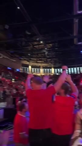 The video shows a lively event, likely a sporting match or concert, in Oslo, Norway. The primary subjects are the enthusiastic crowd and individuals, many wearing red clothing. Large screens are visible, displaying a soccer game with animated graphics. The atmosphere appears celebratory and energetic, with people raising their arms, cheering, and some even dancing. The lighting suggests it's an indoor venue, possibly an arena or stadium, with overhead lights and illuminated screens. The visible text on the screens confirms it's a football game, with team names and scores displayed. The emotions conveyed are excitement and joy, suggesting a positive outcome or a thrilling moment in the event.
