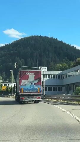 The video shows a view from inside a vehicle, traveling on a road. In the foreground, the dashboard and windshield are visible. The road ahead is paved, with lane markings. A large red truck with the brand "KERN" is in front, also traveling on the road. The truck has a white design featuring a Swiss cross and a fish. To the right of the road, there is a modern, multi-story industrial building with large windows. A sign on the building reads "BURGER GROUP". Behind the building and the road, there is a heavily wooded hill under a clear blue sky with scattered clouds. A tall transmission tower is visible on top of the hill. Traffic lights are present at an intersection. The overall scene is a daytime view of a semi-urban or industrial area surrounded by nature.