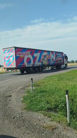 A white semi-truck with an "OZON" logo drives down a two-lane road. The truck is carrying a large, rectangular trailer covered in colorful advertisements. The road is surrounded by green grass and fields under a clear blue sky with a few scattered clouds. The camera angle is low, looking up at the truck as it passes by.