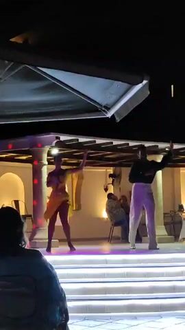 Two dancers perform on a stage at night. The dancer on the left, a woman, wears a bright yellow, fringed dress and heels. She has her arms raised. The dancer on the right, a man, wears a dark shirt and light-colored pants. He has his arms extended to his sides. The stage is lit with white lights on the steps leading up to it, and colored lights illuminate the dancers. In the background, people are seated at tables, and a ceiling fan is visible. The overall environment appears to be an outdoor entertainment area.