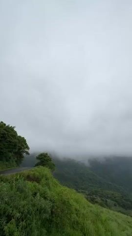 The video showcases a panoramic view of a mountainous landscape under a cloudy sky. The foreground features a grassy hillside, with green vegetation covering the slopes. To the left, a road is visible, lined with trees. The midground reveals a valley with rolling hills covered in grass and scattered trees. A prominent mountain peak is partially obscured by low-hanging clouds or fog. In the background, more distant mountain ranges fade into the horizon under the overcast sky. The overall scene is serene and depicts a natural landscape on a cloudy day.