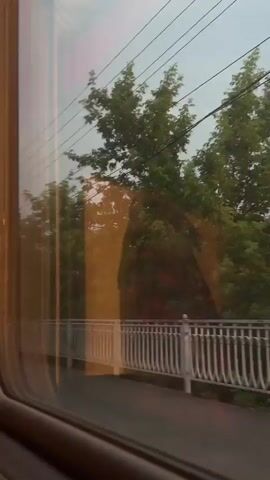 The video shows a view from inside a moving vehicle, looking out a window. The scene outside consists of a railway track with overhead wires and a utility pole. There is a white railing along a sidewalk or platform. Trees and buildings are visible in the background. The sky is overcast and appears to be either early morning or late evening. There are no people or animals visible in the video. The camera appears to be moving forward, as evidenced by the changing perspective of the scenery.