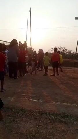 A group of people, mostly children and some adults, are gathered outdoors on a dirt surface in front of a volleyball net. The sun is low in the sky, casting a warm light and creating lens flare. The individuals are casually dressed in t-shirts and shorts. Some are standing, while others appear to be in motion, possibly participating in or observing a game. A few children are walking or running on the court. The environment appears to be a field or open space with some vegetation in the background. A volleyball net is set up, supported by poles. The overall scene suggests a recreational activity taking place in the late afternoon or early evening.