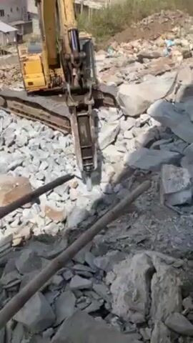 The video shows a yellow excavator breaking up a pile of gray rocks with a jackhammer attachment. The excavator is positioned on top of the rock pile, which slopes downward towards a neighborhood with houses visible in the background. The camera is positioned in front of the excavator, with a few wooden poles visible in the foreground. The excavator's jackhammer is actively hammering into the rocks, creating dust and debris. The setting appears to be a construction or demolition site in a residential area.