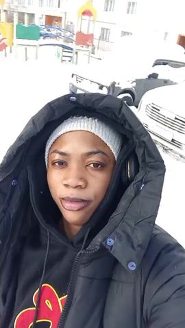 A person wearing a black hooded puffer jacket and gray knit cap is filming a selfie video in a snowy outdoor environment. There are several cars parked nearby covered in snow. Buildings are visible in the background. The person is looking at the camera and makes different facial expressions including sticking their tongue out, and puckering their lips.