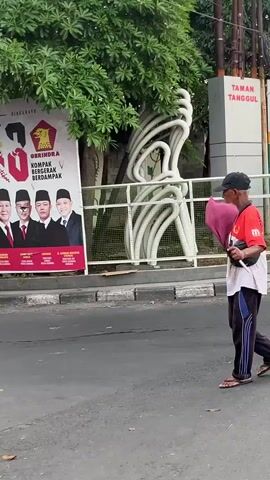 A man walks from right to left across a street carrying a pink bouquet of flowers. He is wearing a red t-shirt, dark pants, and sandals. A large campaign poster for the Indonesian Gerindra political party is visible on the left. The poster features an image of Joko Widodo and other political figures. A white sculpture, resembling intertwined pipes, is located behind a white railing. The man is walking on a paved street, and there is greenery from a tree above. A sign that reads "TAMAN TANGGUL" is visible in the background.