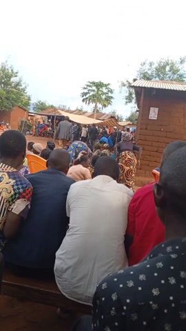The video captures a gathering of people in an outdoor setting, possibly a village or rural area. Most attendees are seated, facing a man dressed in military attire who appears to be speaking to the crowd. There are several houses visible, made of brick and covered with tin roofs. A temporary canopy provides shade for some of the people gathered near the speaking man. The sky is overcast.