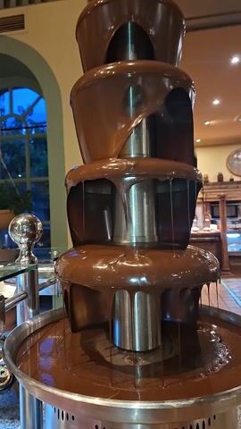 The video shows a chocolate fountain in a breakfast or dessert buffet setup. The fountain has multiple tiers and is actively flowing with chocolate. In the foreground, there's a tray of what appears to be pink marshmallows, possibly strawberry flavored. To the right, there are various pastries laid out on trays in a display case, with a range of shapes and toppings. The background shows elements of a dining area or restaurant, including a window letting in natural light and a serving counter. The focus is primarily on the chocolate fountain and the selection of desserts available.