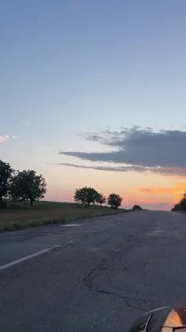The video captures a view from a car driving down a cracked asphalt road at sunset. The sky is a gradient of pale blue to soft orange, with wispy clouds. On the left side of the road, there are fields of dry grass and a few scattered trees. On the right side, more trees line the road, creating a sense of depth. The sun appears to be setting just beyond the horizon, casting a warm glow on the scene. The camera appears to be mounted or held at a low angle, possibly from the dashboard or windshield, as a portion of the car's hood or bumper is visible in the foreground. The overall impression is one of a serene, rural landscape at dusk.