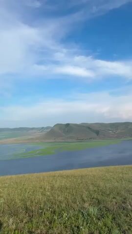 The video shows a panoramic view of a vast river delta with lush green vegetation interspersed with dark blue water under a bright blue sky with scattered white clouds. The foreground consists of a grassy hill. The camera pans slowly across the landscape, offering a wide vista of the natural environment. There are no people or man-made objects visible in the frame. The scene appears to be a serene and expansive natural landscape.