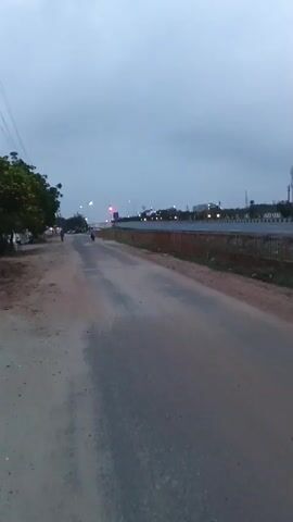 The video captures an outdoor scene on a road. The sky is overcast with a grey hue, suggesting either early morning or late evening. The road surface appears to be a mixture of asphalt and dirt. On the left side of the road, there is some vegetation and a few people are visible walking or riding. Further down the road, there are streetlights and vehicles with their headlights on, indicating low visibility. On the right side, there is a fence, more vegetation, and a few more people walking. The overall atmosphere seems to be one of a busy road in a somewhat rural or suburban setting, possibly experiencing twilight or poor weather conditions. The video is shaky, suggesting it was filmed while moving.