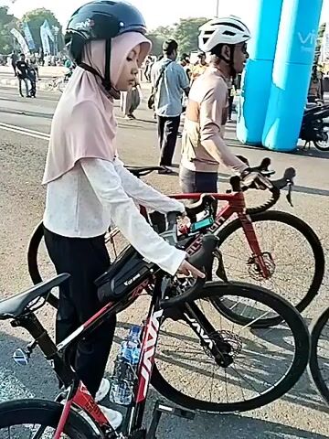 A young woman with light skin, wearing a light pink hijab, a white long-sleeved top, black pants, and a black cycling helmet, is pushing a red and black road bicycle with "JAVA" branding on a paved street. Her bicycle has disc brakes, a water bottle, and a black top-tube bag. To her right, another person in a light brown shirt and white helmet also stands with a bicycle. The camera pans slightly right, revealing numerous other people walking on the street, some casually dressed. Buildings, including one with a "COFFEE" sign, line the right side of the street. Further down the street, white double solid lines are visible, and more pedestrians are walking. In the distance, a fountain or monument is visible. The scene is outdoors on a sunny day.