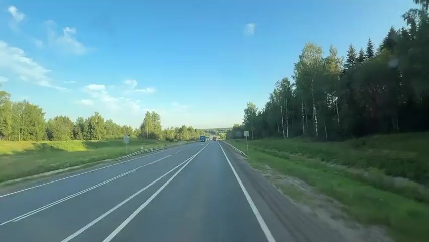 The video shows a first-person perspective driving on a highway through a rural landscape. The road is paved with two lanes in each direction, separated by a double white line. On the left side of the road, there is a grassy shoulder, a metal guardrail, and a line of trees and bushes. On the right side of the road, there is a similar grassy shoulder, guardrail, and a dense forest of coniferous and deciduous trees. The sky is clear blue with a few scattered white clouds. Several trucks are visible on the highway, traveling in the same direction as the camera. The sunlight suggests it is daytime, likely late afternoon or early evening due to the angle of the sun and the shadows. There are no people visible in the video, only vehicles and the natural environment.
