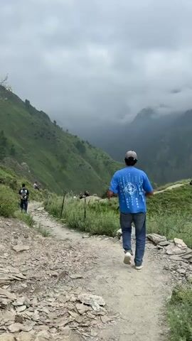A man in a blue t-shirt and jeans walks along a dirt path. The path winds through a mountainous region with green, grassy slopes. In the distance, other people are seen sitting on a hilltop. The sky is cloudy and overcast.
