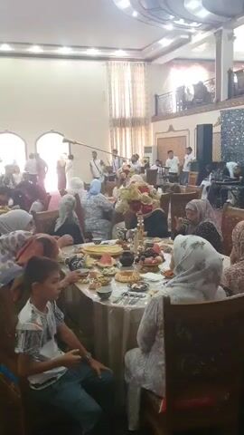 A group of people, likely attending a celebration, are gathered indoors. Many are wearing traditional clothing, with women in headscarves. Several individuals are seated at tables laden with food and drinks. In the background, a band is playing, with a man blowing into a long horn-like instrument and another man playing a drum. Other individuals appear to be standing and observing, some possibly filming the event. The environment suggests a festive occasion, possibly a wedding or similar gathering, with ornate interior decorations and architectural features. The lighting is bright, indicating daytime or well-lit indoor conditions.