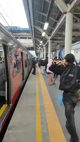 The video shows a train platform with a train stopped at the station. People are standing on the platform, some waiting to board the train, others potentially disembarking. One person in the foreground, wearing a black backpack and dark clothing, walks along the platform away from the camera. The platform has yellow tactile paving along the edge and a covered overhead structure with lighting. The train appears to be red and silver, with its doors open. The overall environment suggests a busy public transportation hub.