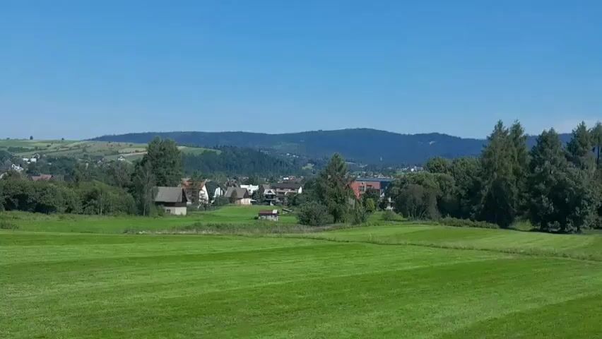 The video shows a panoramic view of a rural landscape under a clear blue sky. In the foreground, there is a neatly mowed green field. In the middle ground, a village is nestled among trees, with houses and some buildings visible. The houses appear to be traditional with pitched roofs. Further in the background, rolling hills and mountains covered in dense green forests rise against the horizon. The scene is bathed in sunlight, suggesting a bright, clear day. The camera is static, offering a continuous, unedited view of the scenery.