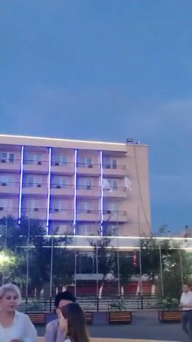 The video shows a scene outdoors at dusk. The sky is a deep blue, indicating twilight. In the background, a multi-story building with several floors is visible. It appears to be a hotel or a similar establishment, adorned with vertical blue lights along its facade. The building has rows of windows and balconies.

In front of the building, there is a street or public area with trees and planters. Several people are visible in the foreground, blurred and out of focus, suggesting they are either moving or the camera is focused on the background. One person in the mid-ground is clearly visible, wearing a white shirt and dark pants, standing and looking towards the right. Another person, a woman in a black outfit, is standing near him, also looking to the right.

The most striking element of the video is a pair of white doves that appear to be flying in the sky near the building. They are the primary focus of the action, as they are the only clearly moving subjects. Their flight suggests a release or a symbolic gesture.

The overall atmosphere is peaceful and perhaps celebratory, given the presence of the doves and the decorative lighting on the building. The time of day suggests an evening event. There are no visible text elements within the video frame. The location context of Baikonur, Kazakhstan, is provided, but not directly discernible from the visual cues in the clip alone.