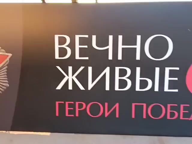 The video shows a wall display with large replicas of Soviet-era medals. The first medal visible is a large, ornate star-shaped medal with a red star in the center and a depiction of what appears to be a military figure in the middle. To the left of this, another medal is partially visible. To the right, the inscription "ВЕЧНЫ ЖИВЫЕ ГЕРОИ" is displayed in large white and red letters. Further to the right, two more medals are prominently featured: one is a star-shaped medal with a depiction of a building inside, and the other is a round medal with a profile of a man's head in the center, which is labeled "ЛЕНИН". The background of the display is dark gray with diagonal striped patterns. The lighting suggests the display is indoors, possibly in a public space like a museum or exhibition hall.