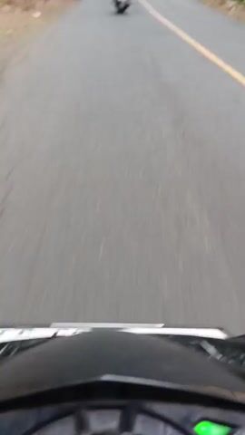 The video shows a first-person perspective of a person riding a motorcycle on a paved road. The road is lined with trees on both sides, and there are some buildings visible in the distance. The rider is following another motorcycle, which is visible ahead. The weather appears to be overcast. The road has some cracks and potholes.