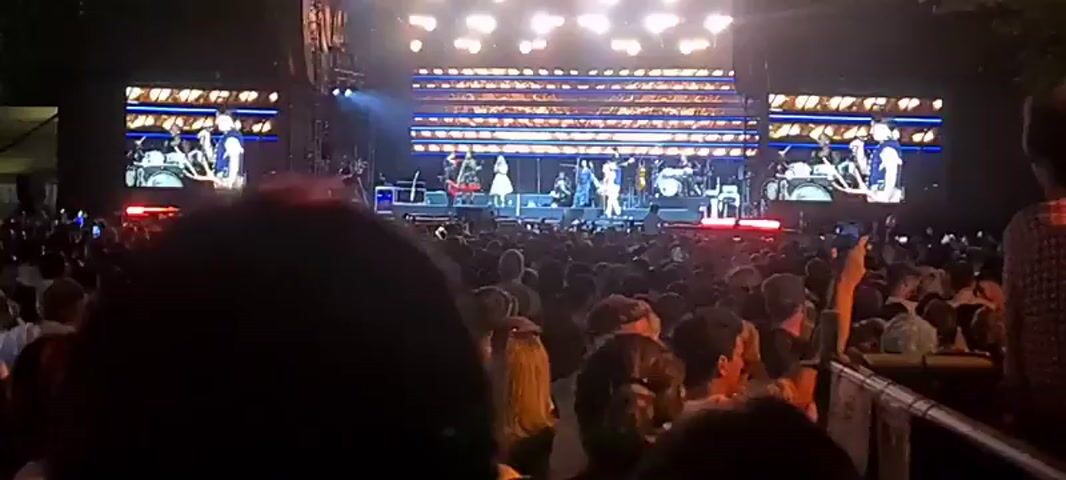 This is a wide shot of a live concert. A band is performing on a brightly lit stage. The band consists of several musicians, including a guitarist on the left, a drummer in the back, and vocalists in the center. Two large screens on either side of the stage display live feeds of the performers. A large crowd of concertgoers is visible in the foreground, facing the stage. The environment appears to be outdoors at night, with trees visible in the background.