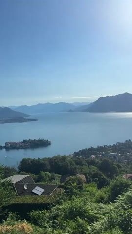 The video is a vertical, first-person perspective video taken from a high vantage point overlooking a large body of water, likely a lake, with mountains in the background. The sun is high in the sky, casting a bright glare on the water. In the foreground, there is lush green vegetation, including trees and ferns, with a house and a satellite dish partially visible on the left. In the midground, there is a town or village nestled along the shoreline, with buildings and trees. There is a small island with buildings on it in the distance. The water is calm with some small ripples and a few boats are visible on the water. The sky is clear and blue. The video pans slowly to the right, revealing more of the coastline and the surrounding mountains. The overall impression is one of a scenic, peaceful landscape.