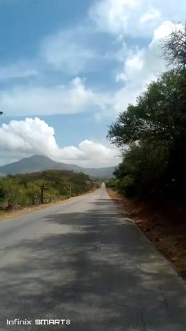 The video shows a straight, asphalt road stretching into the distance. The road is flanked by green vegetation on both sides. On the left, there is sparse, low-lying brush and utility poles. On the right, there are taller trees.  A mountain range is visible in the background, partially obscured by fluffy white clouds in a blue sky.  The road appears to be empty except for a small vehicle in the far distance.  The video is taken from a low vantage point, and the words "Infinix SMART 8" are visible in the lower left corner of the screen.