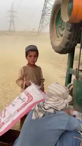 The video captures a dusty rural scene in 'Alaqadari Daman, Afghanistan, featuring a young boy and an adult engaged in agricultural work. A young boy, wearing a traditional dark cap and a light brown striped tunic, stands with a serious expression. He holds a large white and pink patterned sack. Next to him, an adult, dressed in a light blue garment and with a striped cloth covering their head, is partially visible, appearing to be seated or stooping while working with a green and orange agricultural machine, possibly a thresher, of which a large wheel is prominently seen. The air is thick with dust and chaff, indicative of an ongoing harvesting or processing activity. In the hazy background, several power line towers (pylons) are visible. The setting suggests a day of labor in a dry, agricultural field under a diffused sky. The text on the sack is not clearly legible.