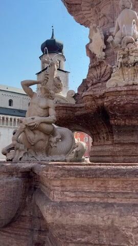 The video showcases a large, ornate fountain with classical statues in a sunlit outdoor setting. The fountain, made of what appears to be stone with pinkish and tan hues, is the central focus. It features multiple tiers with figures that seem to depict mythological or allegorical characters.

One prominent statue on the left shows a muscular male figure wrestling with a sea creature, possibly a dragon or serpent, which is coiled around him. Water is spraying from the mouth of this creature, contributing to the fountain's active display. Higher up on the fountain, another statue of a child or cherub is visible, with water cascading around it. Further to the right, another female figure stands, also appearing to be part of the fountain's sculptural program. The water from the fountain creates a dynamic effect, with droplets catching the sunlight.

In the background, a tall, steeple-topped building, likely a church or cathedral, is visible under a clear blue sky with scattered white clouds. The architecture of this building suggests an old European city. The overall scene evokes a sense of grandeur and historical significance. The bright sunlight indicates it is daytime, likely late morning or afternoon, and the clear sky suggests good weather. The presence of the fountain and the historical architecture points to a public square or plaza in a European city. While no visible text is present in the video, the style of the statues and architecture are characteristic of Italian Renaissance or Baroque art, placing the scene in Italy, specifically Trento as indicated by the context. The activity is simply the functioning of a public fountain, creating a lively and picturesque atmosphere.