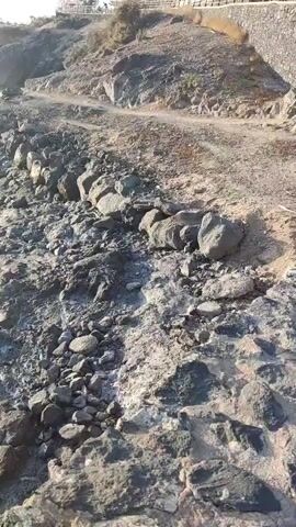The video begins with a close-up of a rocky coastline. The terrain is uneven and covered in various sizes of stones and boulders. A path or dirt track is visible in the upper right portion of the frame, winding along the top of a rocky incline. The camera then pans to the left, revealing the ocean meeting the rocky shore. Small waves are gently lapping against the rocks. As the camera continues to pan, the sun becomes visible in the sky, casting a bright glare on the water's surface. The sun is low in the sky, suggesting it may be sunrise or sunset. In the distance, a breakwater or a line of submerged rocks stretches across the water. There are no people clearly visible in the initial frames, but as the camera pans, a figure can be briefly glimpsed standing on the rocks near the water's edge in the distance. The overall environment is natural and somewhat rugged, with a clear sky and the ocean as the dominant features.