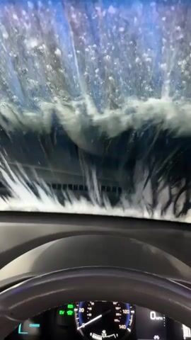 The video shows the view from inside a car as it goes through a car wash. Large amounts of water and soap are being sprayed onto the windshield. The car's dashboard, steering wheel, and speedometer are visible in the foreground. The speedometer reads 0 MPH.
