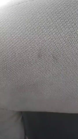 The video shows a close-up view of a textured fabric surface, possibly upholstery. The lighting is diffused, and the surface appears to be light in color, likely gray or off-white, with a woven pattern. There are some darker smudges or stains visible on the fabric. The camera is positioned at an angle, suggesting it is either being held or is attached to an object. There is no discernible action, interaction, or identifiable environment beyond the fabric itself.