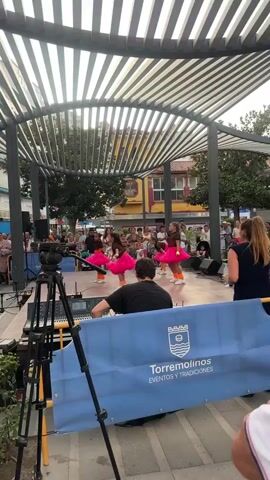 A video captures a group of young girls performing a dance routine on an outdoor stage. The girls are dressed in matching outfits featuring brown tops and vibrant pink tutus. They are all wearing white sneakers and orange leg warmers. Each dancer is holding a yellow object, possibly a ball or a prop. The stage is covered by a slatted roof structure, with trees and buildings visible in the background. A crowd of spectators is gathered to watch the performance. In the foreground, a sound mixing board and a video camera on a tripod are positioned. A person is seated behind the sound board, presumably operating it. A blue banner with the text "TORREMOLINOS EVENTOS Y TRADICIONES" and a crest is visible at the bottom of the frame. The lighting suggests it is daytime, likely overcast or in the shade.