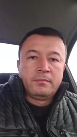 A man with dark hair and brown eyes, wearing a black quilted jacket over a dark sweater, sits in the driver's seat of a car. He looks directly at the camera. The car is moving, as the background outside the window changes. The sky is overcast. Other cars and buildings are visible through the window.
