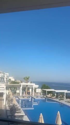 A resort area with a swimming pool and ocean view. The pool is blue and surrounded by lounge chairs. There are umbrellas on the pool deck. In the background, there is a white building with palm trees. The ocean is calm and blue, with a clear sky above. The video pans across the scene, showing the different elements of the resort. There are no people visible in the video.