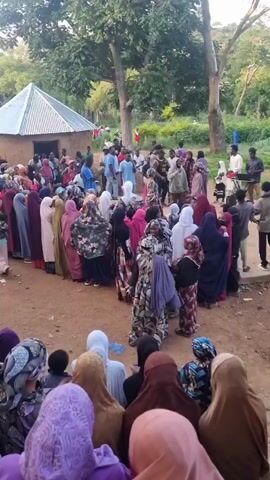 A crowd of people is gathered outdoors. A group of men and women are standing in a line. The people are dressed in a variety of clothing, including headscarves and traditional attire. There are trees and a small building in the background. The ground is dirt. Some people are looking at a table set up in the middle of the gathering. The overall impression is of a community event or gathering.