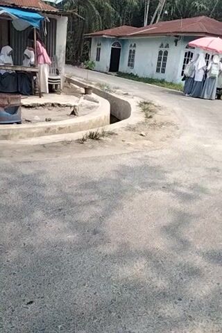 The video is a vertical-oriented, low-resolution recording of an outdoor scene, likely a street or pathway next to a building. In the foreground, there is a paved surface with scattered debris and shadows cast by trees. To the left, a light-colored, single-story building with a red-tiled roof is visible. The building has multiple windows and a dark-colored door. In front of the building, there is a collection of what appears to be folded clothes or fabric stacked on a cart or stand, covered by a pink umbrella. Several individuals, dressed in what looks like school uniforms with headscarves, are gathered near the cart. There is also a motorcycle parked near the group. In the lower left corner, there are more items of clothing laid out. The environment appears to be warm and sunny, with vegetation visible around the building. The video is shaky, suggesting it was handheld.