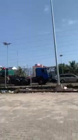 The video begins with a view of a flatbed truck carrying an excavator. The camera pans to the right, revealing a paved area with parked vehicles and a temporary barrier. Two women walk into the frame, followed by two young men, one in a school uniform. The camera then pans back to the right, showing more of the paved area and the barrier. The overall setting appears to be an outdoor area with some construction equipment and activity.
