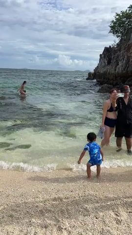 A group of people are enjoying a beach vacation in Tambon Phru Nai, Thailand. The scene takes place on a sandy beach with clear, shallow water lapping at the shore. In the foreground, a young child in a blue and white swimsuit stands facing the water, his arms outstretched as if reaching for something. Behind him, a couple poses for a selfie with a smartphone. The woman is wearing a navy blue bikini, and the man is dressed in a black open-collared shirt and shorts, with a baseball cap. Further out in the water, another person, likely a woman, is swimming. To the right of the frame, a rocky cliff face covered in lush green foliage juts out into the sea. The sky is partly cloudy, suggesting it is daytime, possibly late morning or early afternoon. The overall atmosphere appears relaxed and happy, indicative of a pleasant day at the beach. No visible text is present in the media.