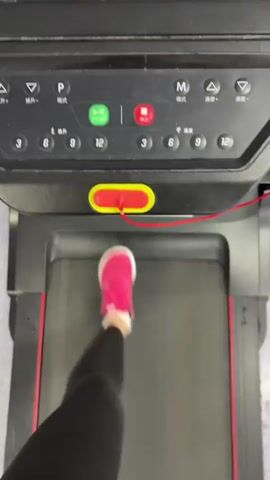 From a high-angle perspective, the video captures a person walking on a treadmill. The person is wearing black leggings and bright pink sneakers. The treadmill's control panel is visible, featuring an array of buttons and a yellow safety stop with a red cord attached.  On top of the control panel, there are several small objects, including a pink AirPod case, a small white key chain, and a red rectangular object.  The treadmill itself has a black and red frame with "GO RUNNING" printed on the side. The background is a light-colored floor.
