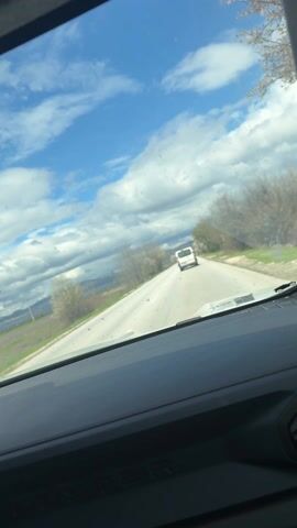 The view from inside a car shows a bright, sunny day with scattered white clouds against a blue sky. The car is traveling on a grey, paved road with a white van driving ahead. On either side of the road are green fields, small trees, and shrubbery. In the distance, hills can be seen. The windshield of the car is somewhat dirty with spots. The dashboard is visible in the foreground, providing a frame for the outdoor scene.
