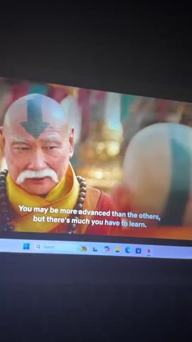A person is filming a laptop screen showing a scene from a TV show or movie. An older man with a bald head with a blue arrow painted on it, a white mustache, and a red and yellow robe speaks. He is wearing a strand of beads as well. The subtitles on the screen read, "You may be more advanced than the others, but there's much you have to learn. And believe it or not." Then the camera angle shows the back of the man's head as he hugs a young boy who also has a bald head with a blue arrow painted on it.